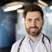 Smiling male doctor with beard and stethoscope standing in modern medical office. Confident physician dressed in white coat ready for patient care and consultation in contemporary healthcare setting.