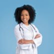 Positive confident young african american curly woman doctor therapist in white coat with crossed arms on chest, isolated on blue background, studio. Medicine service, health care, treatment